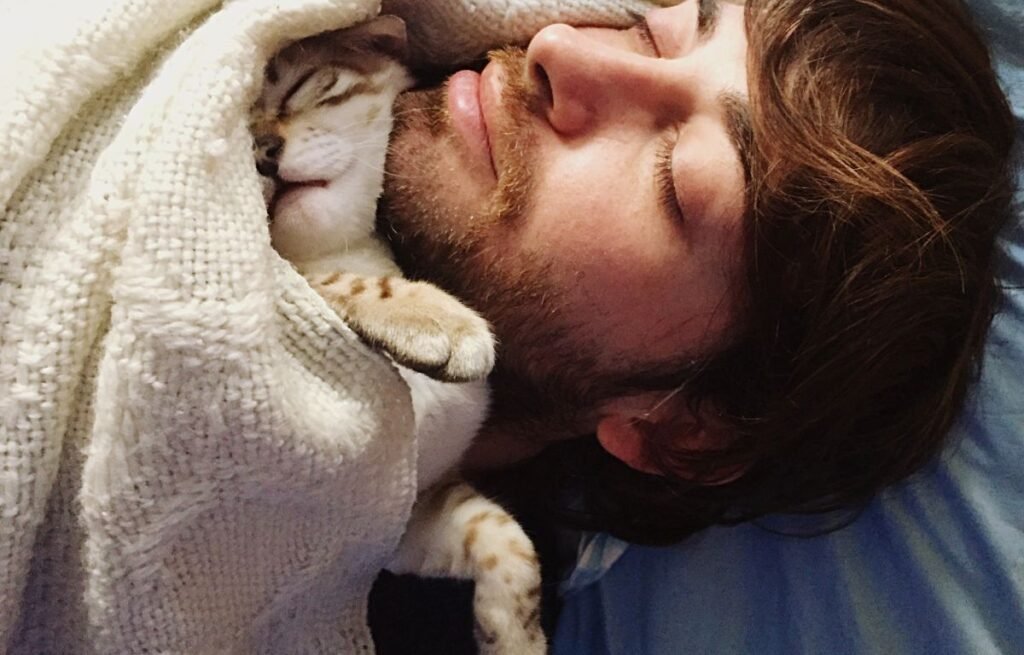 Why Does My Cat Sleep on Me Every Night – A peaceful scene of a cat and its owner sleeping closely together under a blanket, showing trust, warmth, and emotional bonding between cats and humans.